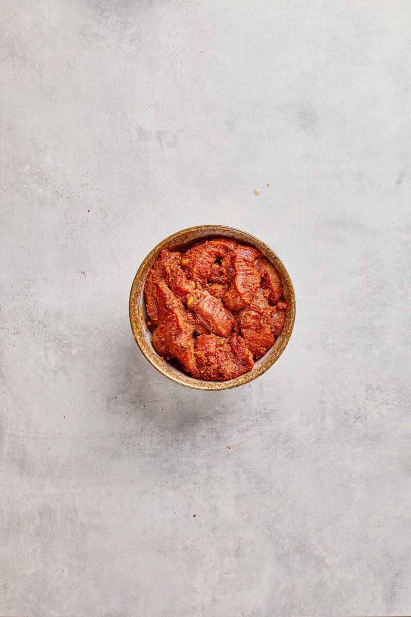 A small bowl filled with seasoned, marinated meat placed on a light gray textured surface.