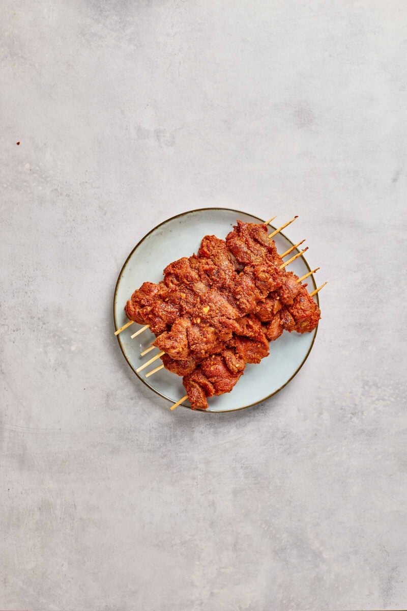 A plate with several raw skewers of marinated grilled meat arranged neatly on a light-colored surface.