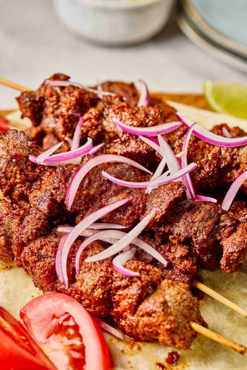 Close-up of spicy grilled suya skewers topped with sliced red onions, served with tomato slices and lime wedges on a flatbread.