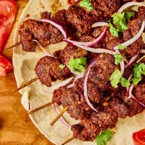 Skewers of spiced grilled beef served on flatbread, garnished with sliced red onions and fresh cilantro, with tomato slices on the side.