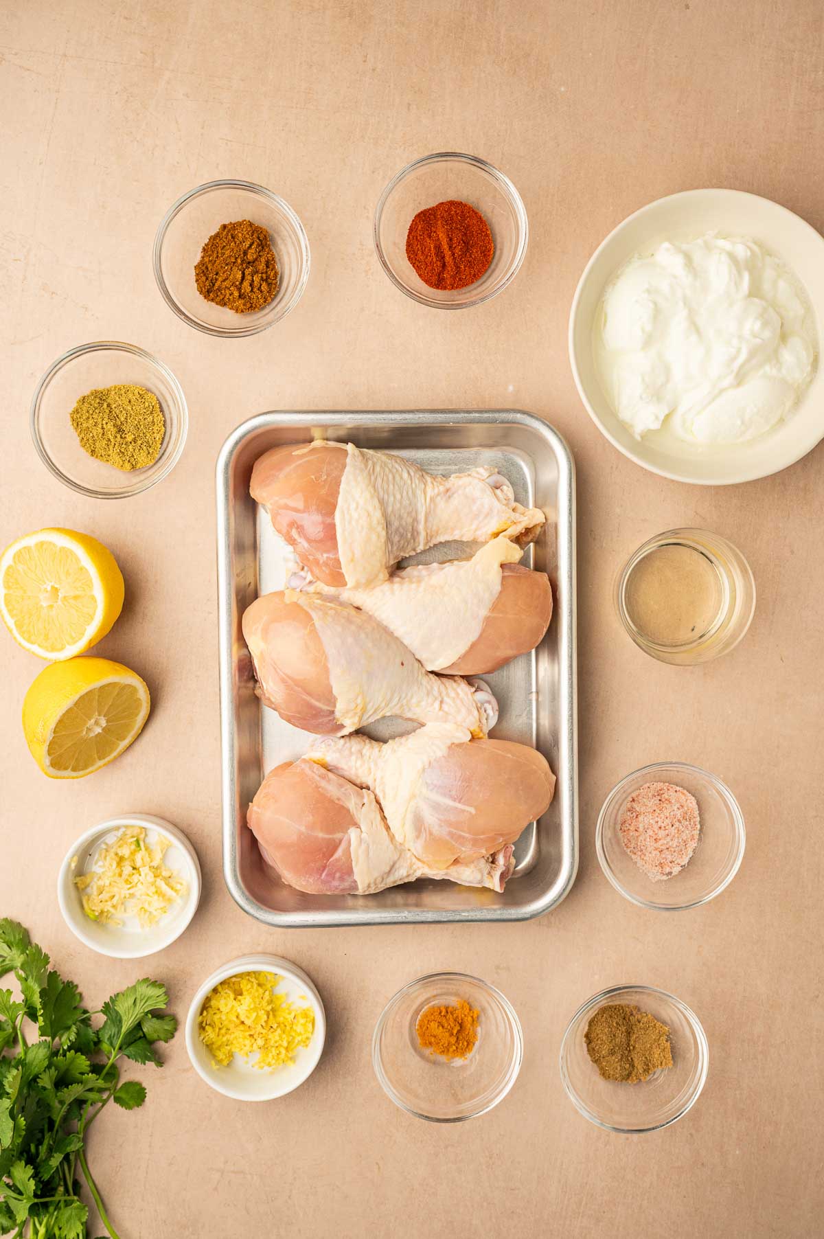 A tray with raw chicken legs is surrounded by small bowls containing various spices, yogurt, lemon halves, minced garlic, ginger, cilantro, and a small glass of liquid.