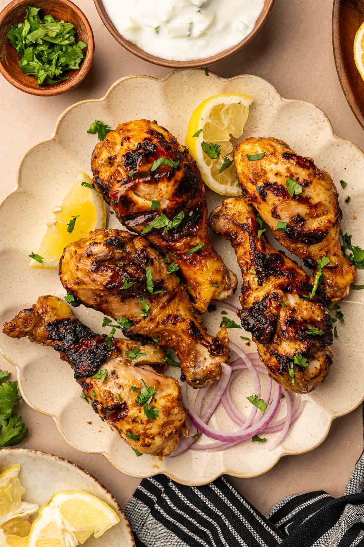 Plate of grilled tandoori chicken drumsticks garnished with chopped herbs, sliced red onions, and lemon wedges, with small bowls of raita and herbs nearby.