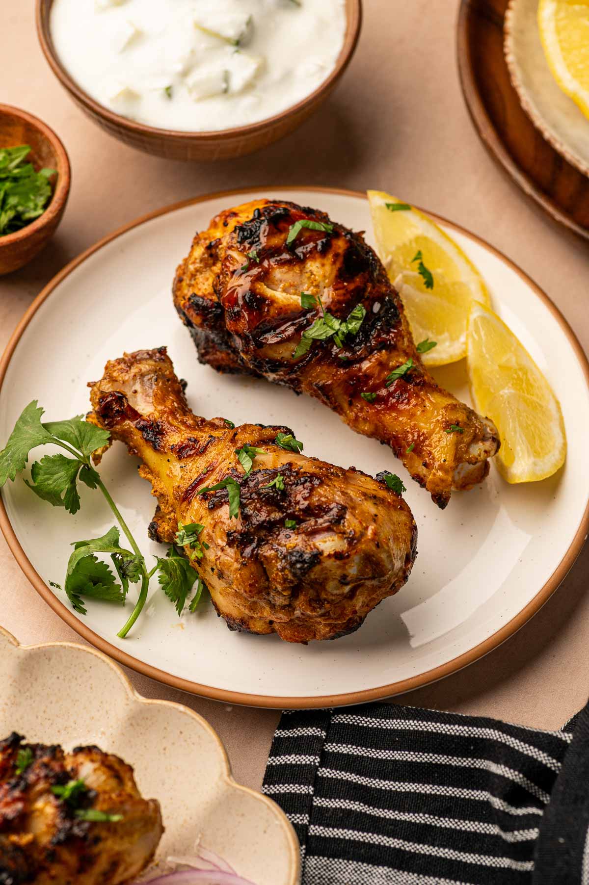 Two grilled tandoori chicken drumsticks garnished with cilantro served on a white plate with lemon wedges; small bowls of yogurt sauce and chopped herbs nearby.
