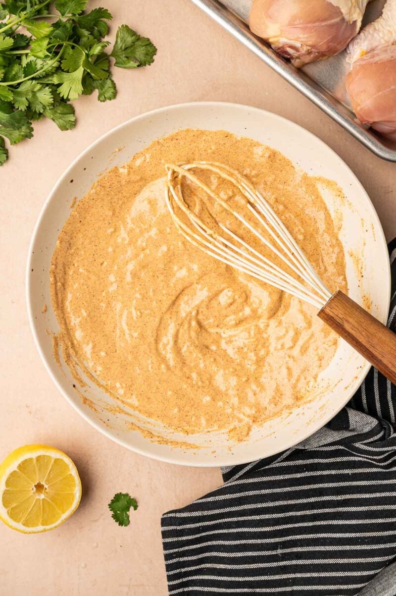 A bowl of tandoori chicken marinade being whisked, surrounded by fresh cilantro, raw chicken pieces on a tray, a striped towel, and a halved lemon on a beige surface.