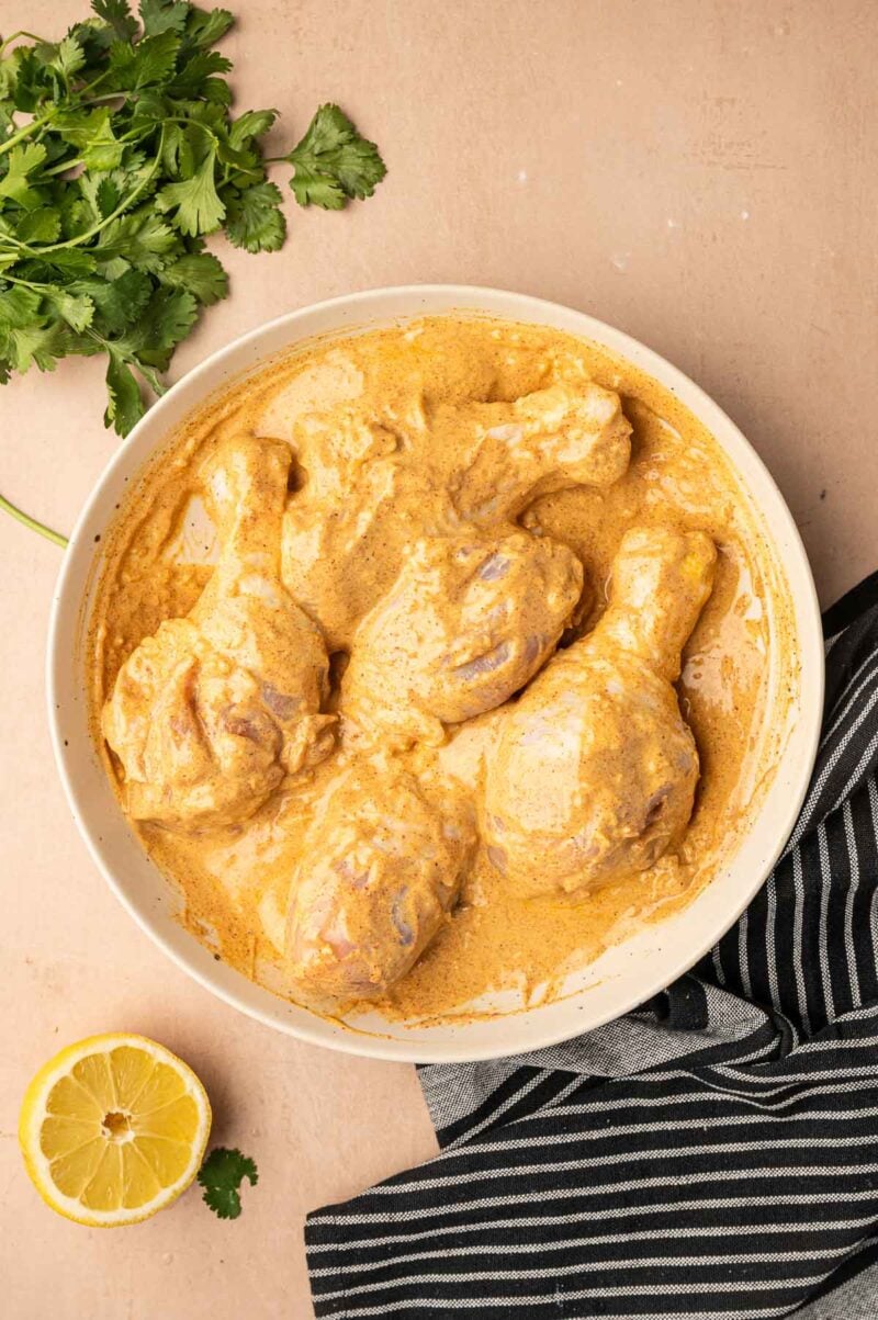 Raw chicken drumsticks coated in a creamy yellow tandoori marinade sit in a round bowl, with a bunch of cilantro, a halved lemon, and a black striped towel nearby.