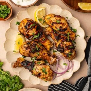 Grilled tandoori chicken drumsticks garnished with herbs and served with lemon wedges, sliced red onion, fresh cilantro, yogurt sauce, and extra lemon on the side.