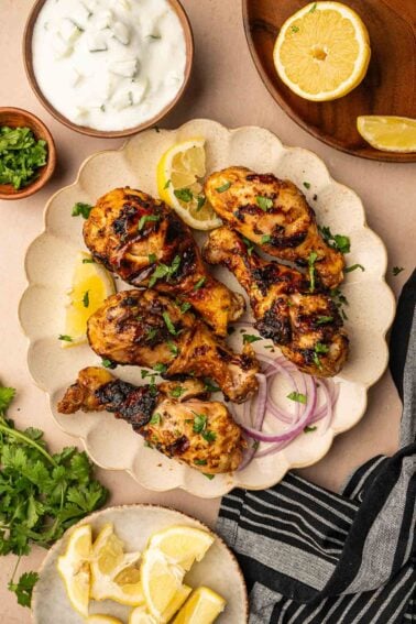 Grilled tandoori chicken drumsticks garnished with herbs and served with lemon wedges, sliced red onion, fresh cilantro, yogurt sauce, and extra lemon on the side.