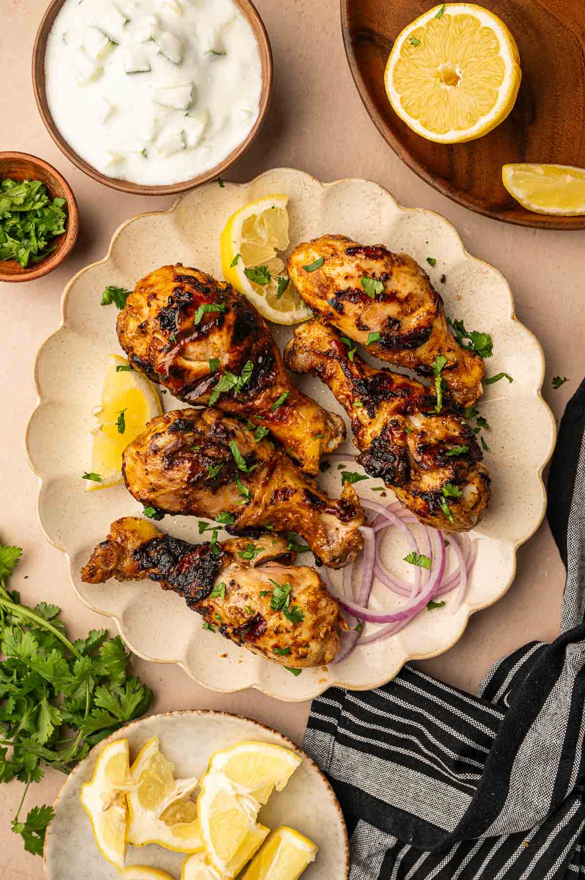 Grilled tandoori chicken drumsticks garnished with herbs and served with lemon wedges, sliced red onion, fresh cilantro, yogurt sauce, and extra lemon on the side.