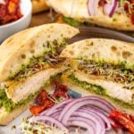 A sliced grilled chicken sandwich with pesto, sprouts, red onion, sun-dried tomatoes, and ciabatta bread is served on a plate with extra onion and sun-dried tomatoes on the side.
