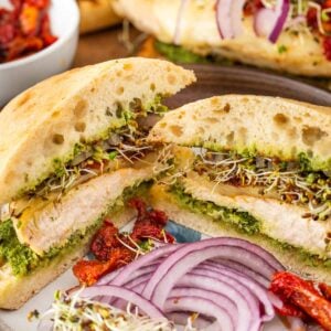 A sliced grilled chicken sandwich with pesto, sprouts, red onion, sun-dried tomatoes, and ciabatta bread is served on a plate with extra onion and sun-dried tomatoes on the side.
