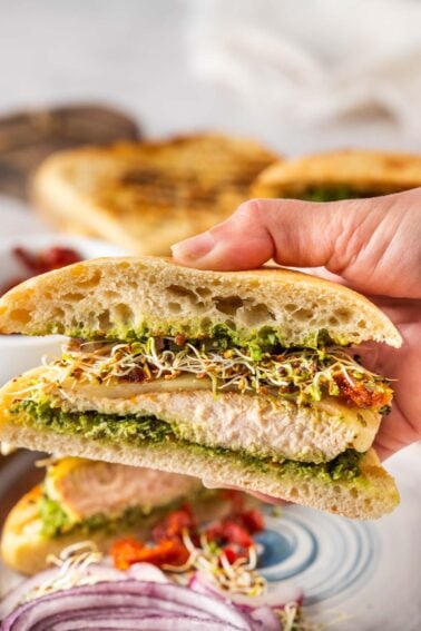 A hand holds a sandwich with focaccia bread, grilled chicken, green pesto, cheese, sprouts, and sun-dried tomatoes. Sliced red onions are visible in the foreground.