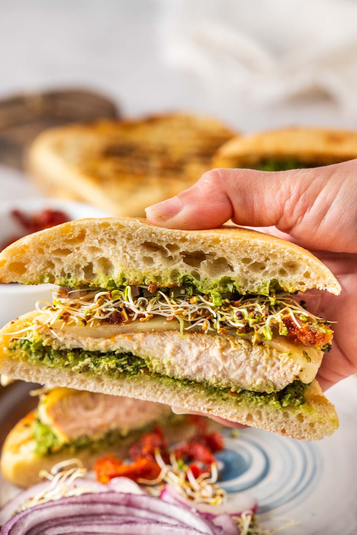 A hand holds a sandwich with focaccia bread, grilled chicken, green pesto, cheese, sprouts, and sun-dried tomatoes. Sliced red onions are visible in the foreground.