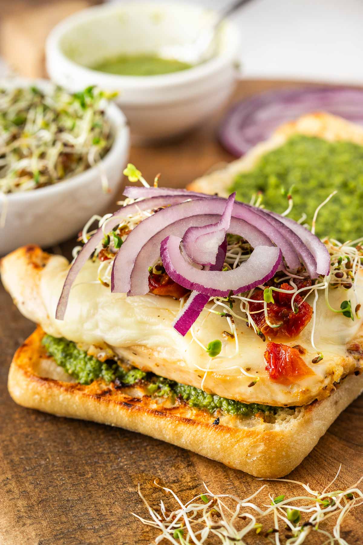 Grilled chicken sandwich on ciabatta bread with pesto, melted cheese, sun-dried tomatoes, alfalfa sprouts, and red onion slices; a bowl of sprouts and sauce in the background.