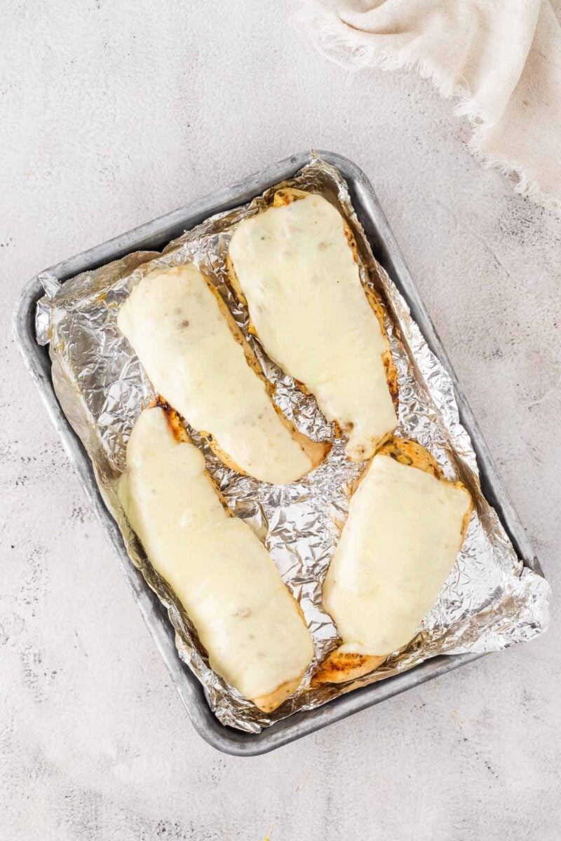 Four pieces of chicken breast topped with melted cheese on a foil-lined baking sheet.