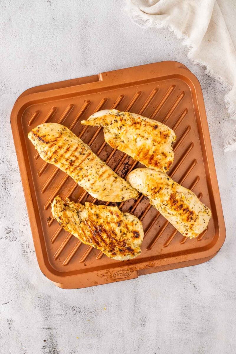 Four grilled chicken breasts on a square copper grill pan, placed on a light surface with a white cloth in the corner.