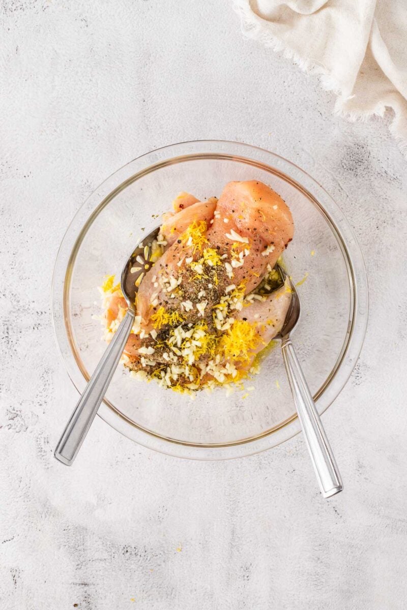 Raw chicken breasts in a glass bowl with grated lemon zest, minced garlic, herbs, pepper, and two metal spoons, ready for marinating on a light surface.