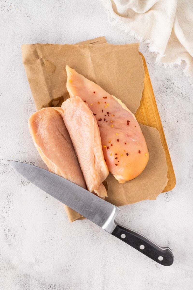 Two raw chicken breasts, being sliced in half for even thickness, one seasoned with red pepper flakes, on brown paper with a kitchen knife beside them on a wooden board.