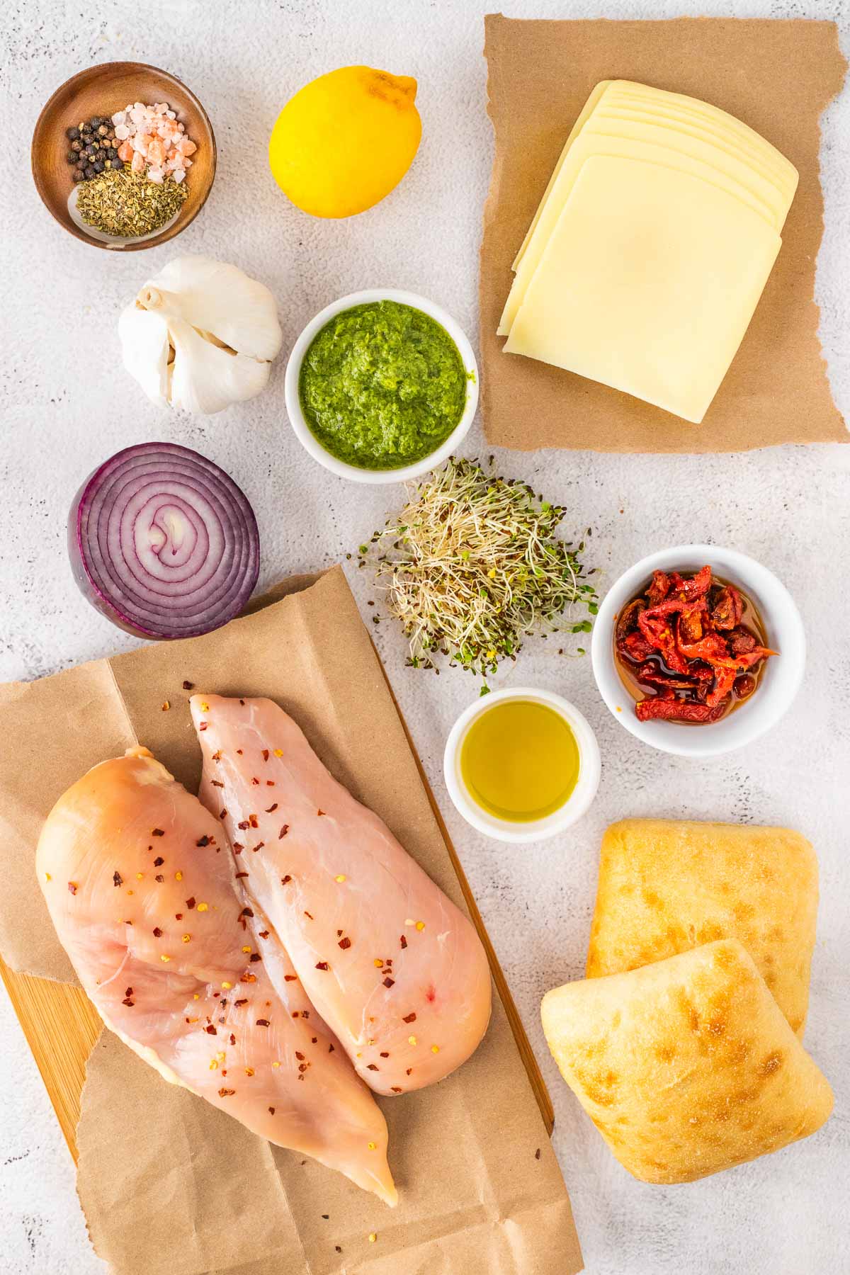 Raw chicken breasts, slices of cheese, red onion, lemon, garlic, pesto, sprouts, sun-dried tomatoes, olive oil, spices, and two pieces of bread arranged on a white surface.