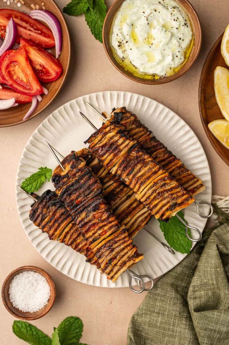 A pile of grilling skewers of layered with tortilla and meat layers, served with tzatziki, lemon wedges, sliced tomatoes, onions, and a bowl of salt.