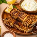 Two grilled potato skewers on a wooden plate with lemon wedges, a bowl of yogurt dip, and a small dish of coarse salt, garnished with mint leaves.