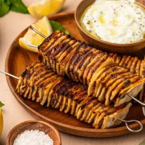 Two grilled potato skewers on a wooden plate with lemon wedges, a bowl of yogurt dip, and a small dish of coarse salt, garnished with mint leaves.