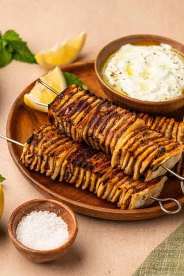 Two grilled potato skewers on a wooden plate with lemon wedges, a bowl of yogurt dip, and a small dish of coarse salt, garnished with mint leaves.