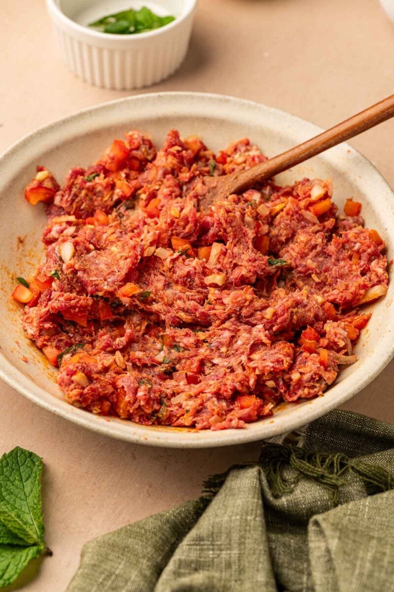 A bowl filled with a mixture of ground lamb, chopped vegetables, and herbs, with a wooden spoon resting in the mixture.