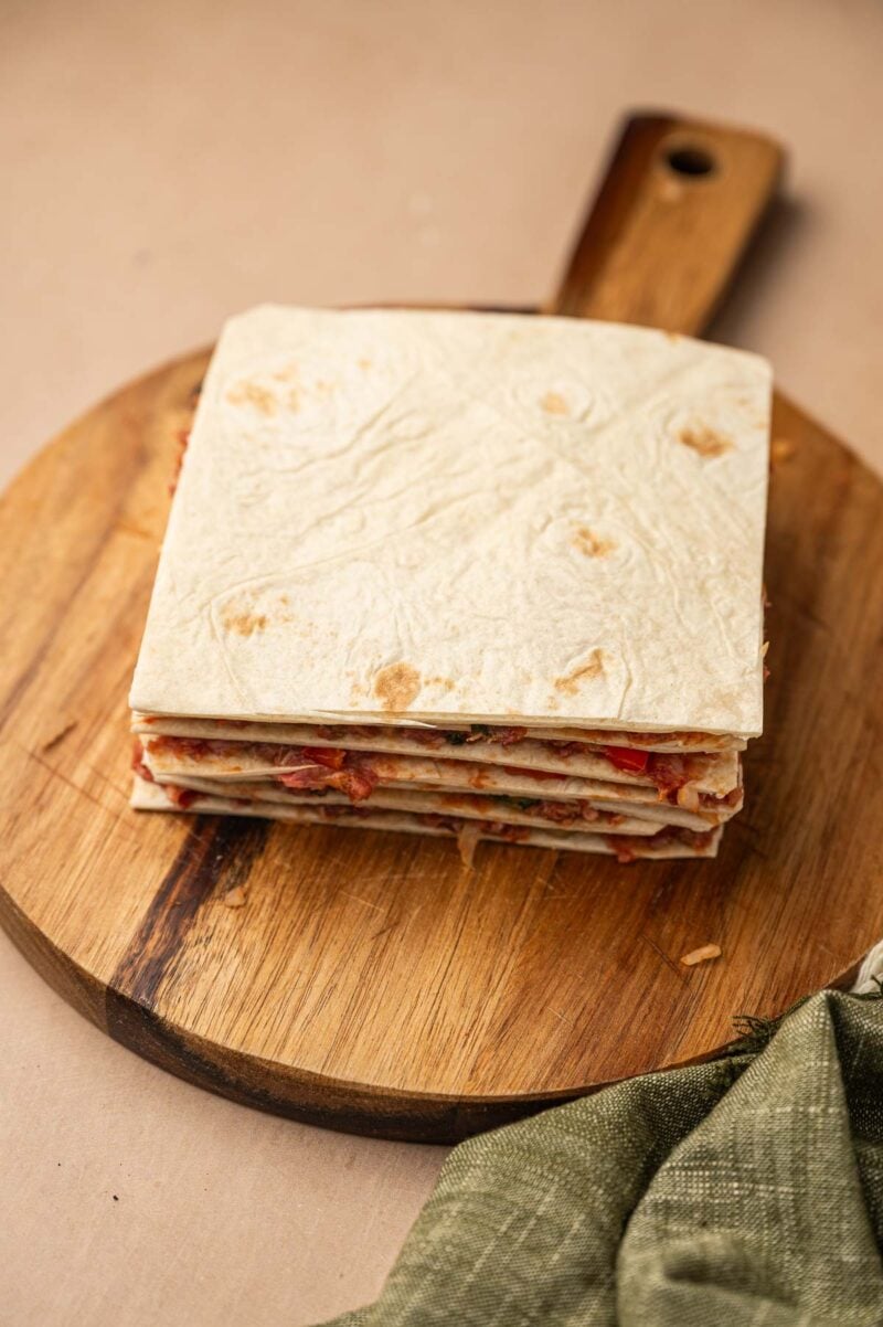 A stack of square tortillas stacked into layers with ground lamb filling sits on a round wooden cutting board, with a green cloth nearby.