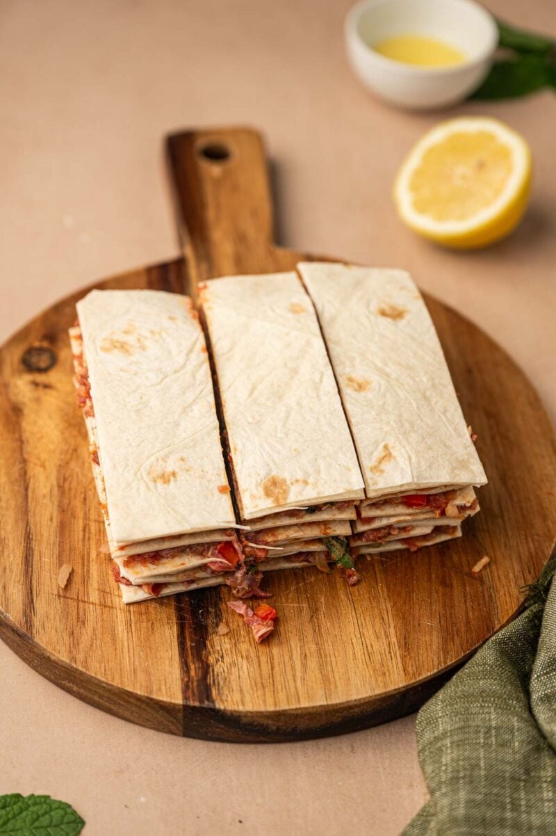 A stack of layered meat filled tortillas cut into strips on a round wooden cutting board, with a lemon half and small bowl in the background.