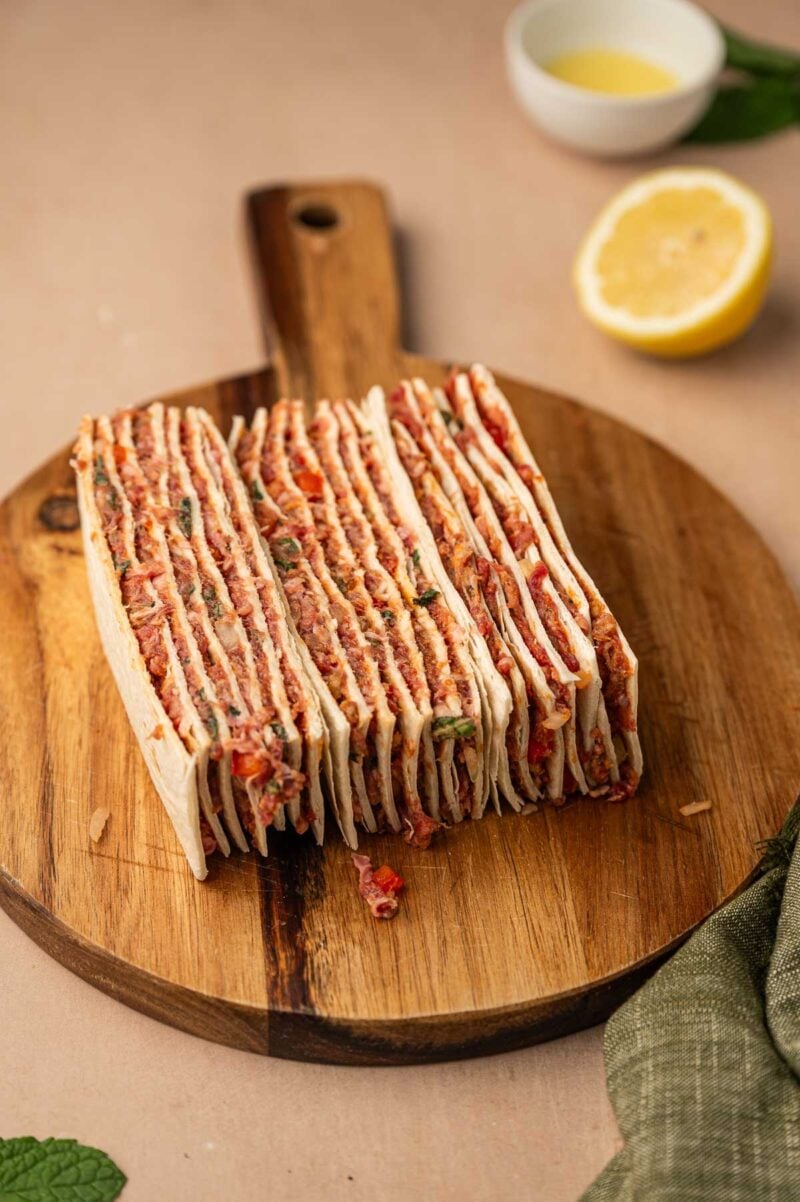 A stack of thin, layered tortillas filled with a minced lamb mixture is arranged on a round wooden cutting board on its side to easily skewer with metal kebobs.