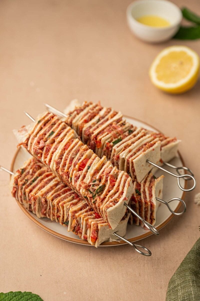 Metal skewers hold stacks of thin, square cuts of tortillas layered with ground lamb filling, arranged on a plate.