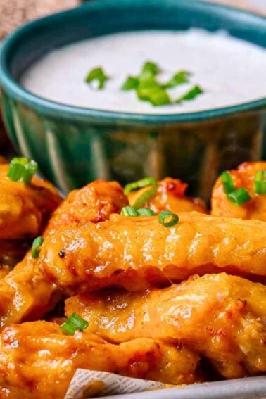 A tray of buffalo chicken wings garnished with chopped green onions, served with a bowl of ranch dip in the background.