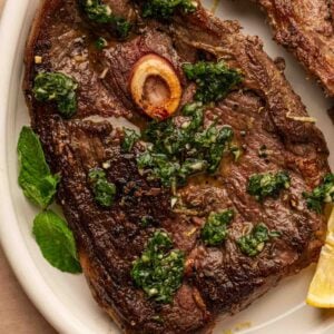 Seared lamb leg steaks with a bone, topped with mint sauce, served with lemon wedges and mint leaves on a white plate.