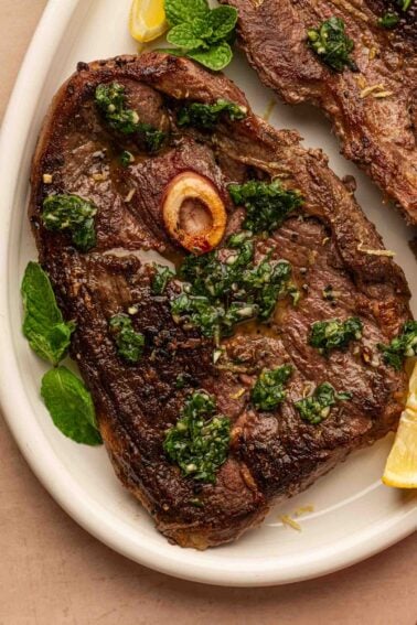 Seared lamb leg steaks with a bone, topped with mint sauce, served with lemon wedges and mint leaves on a white plate.