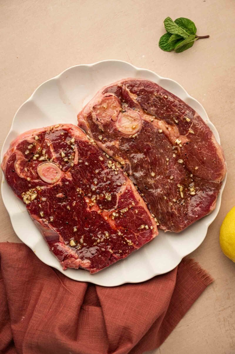 Two raw marinated lamb steaks with visible seasonings on a white plate, placed on a beige surface with a brown cloth, mint leaves, and a lemon nearby.