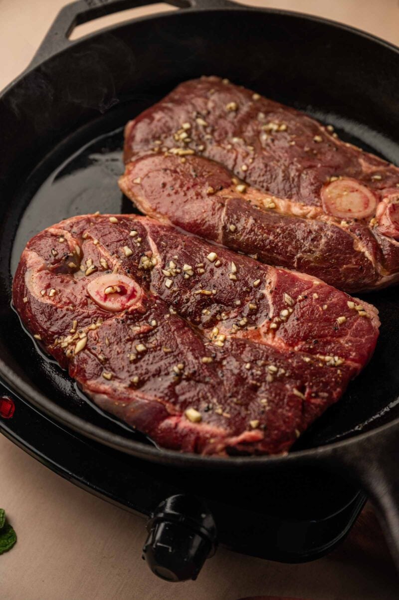 Two seasoned lamb leg steaks searing in a black cast iron skillet.