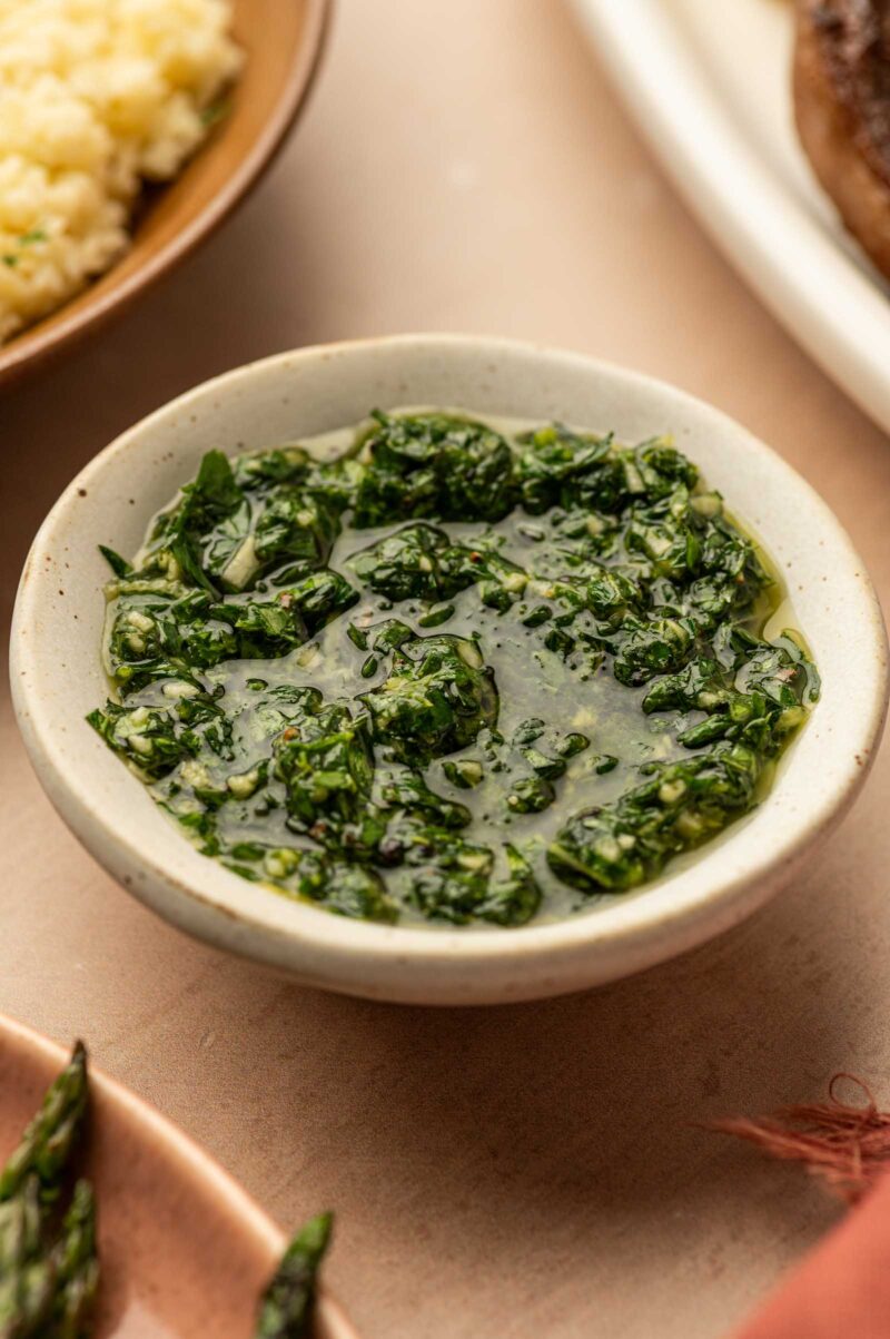 A small bowl filled with green mint gremolata sauce, placed on a light-colored surface with parts of other dishes visible nearby.