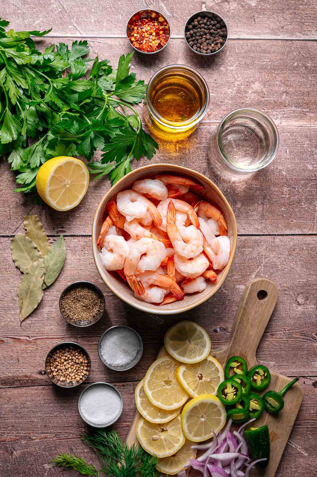 A bowl of shrimp surrounded by fresh parsley, lemon, spices, oil, vinegar, sliced jalapeños, dill, and red onion on a wooden surface.