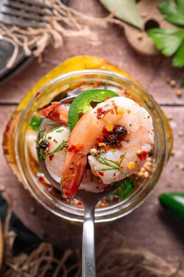 A close-up of a metal spoon holding a pickled shrimp with red pepper flakes, dill, and a slice of jalapeno, with a glass jar of shrimp and spices in the background.