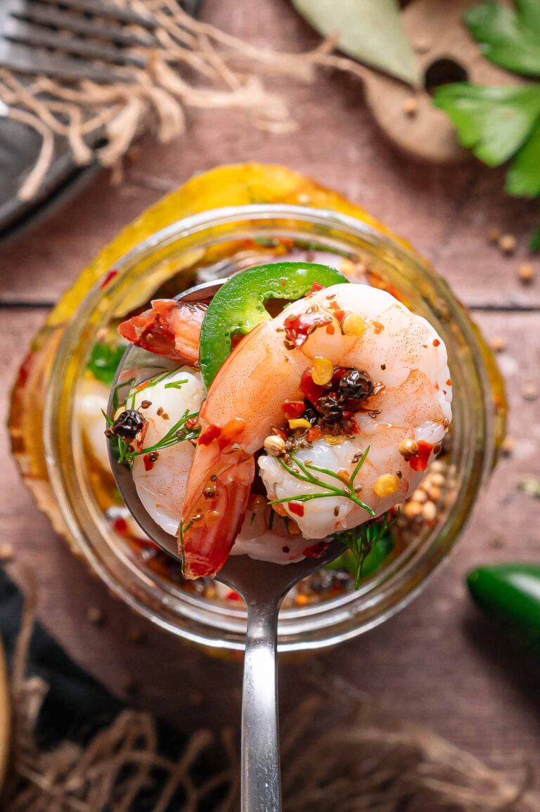 A close-up of a metal spoon holding a pickled shrimp with red pepper flakes, dill, and a slice of jalapeno, with a glass jar of shrimp and spices in the background.