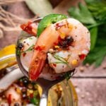 A close-up of a spoon holding a pickled shrimp garnished with herbs, chili flakes, and peppercorns, with lemon and parsley in the background.