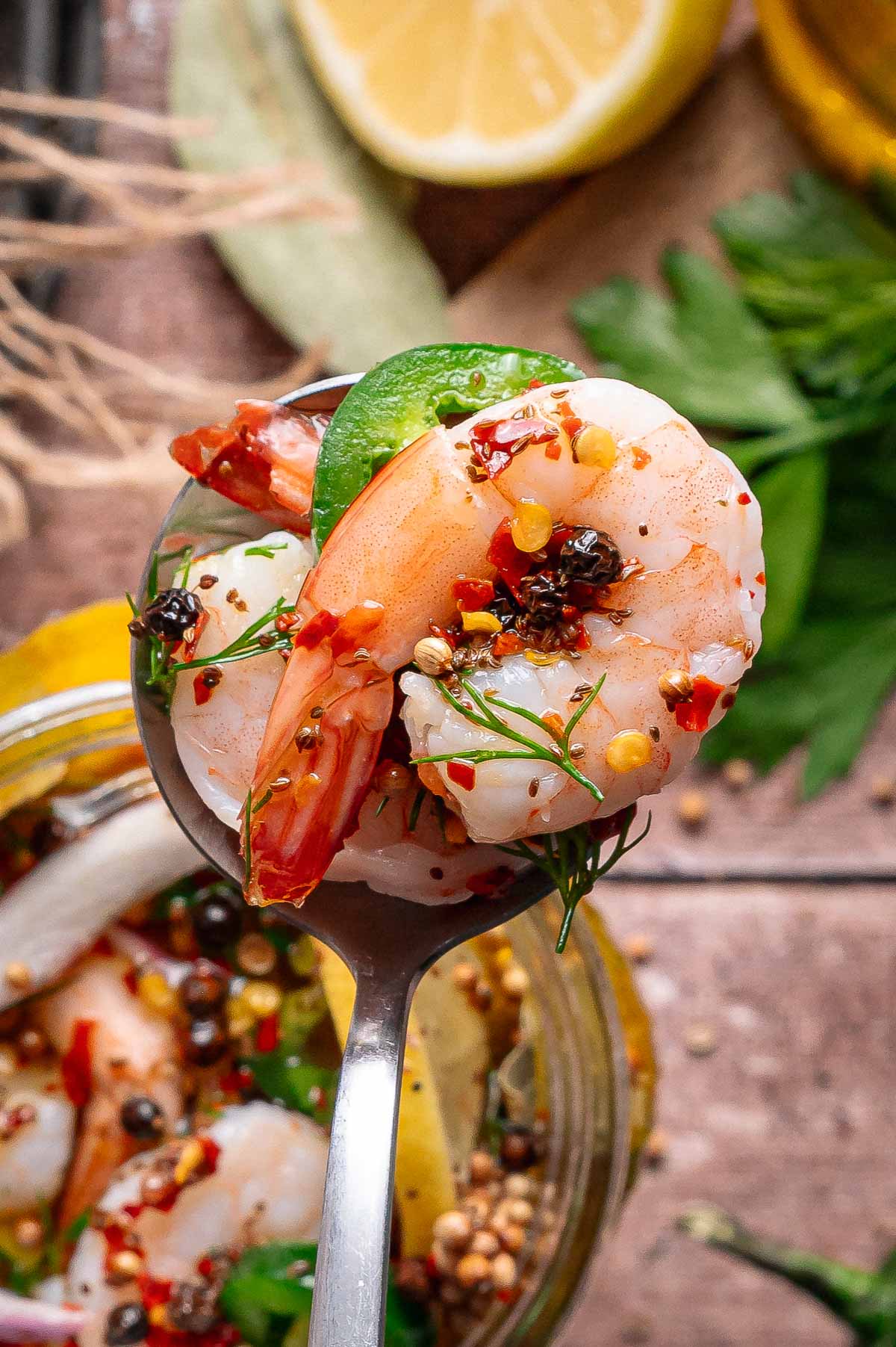 A close-up of a spoon holding a pickled shrimp garnished with herbs, chili flakes, and peppercorns, with lemon and parsley in the background.