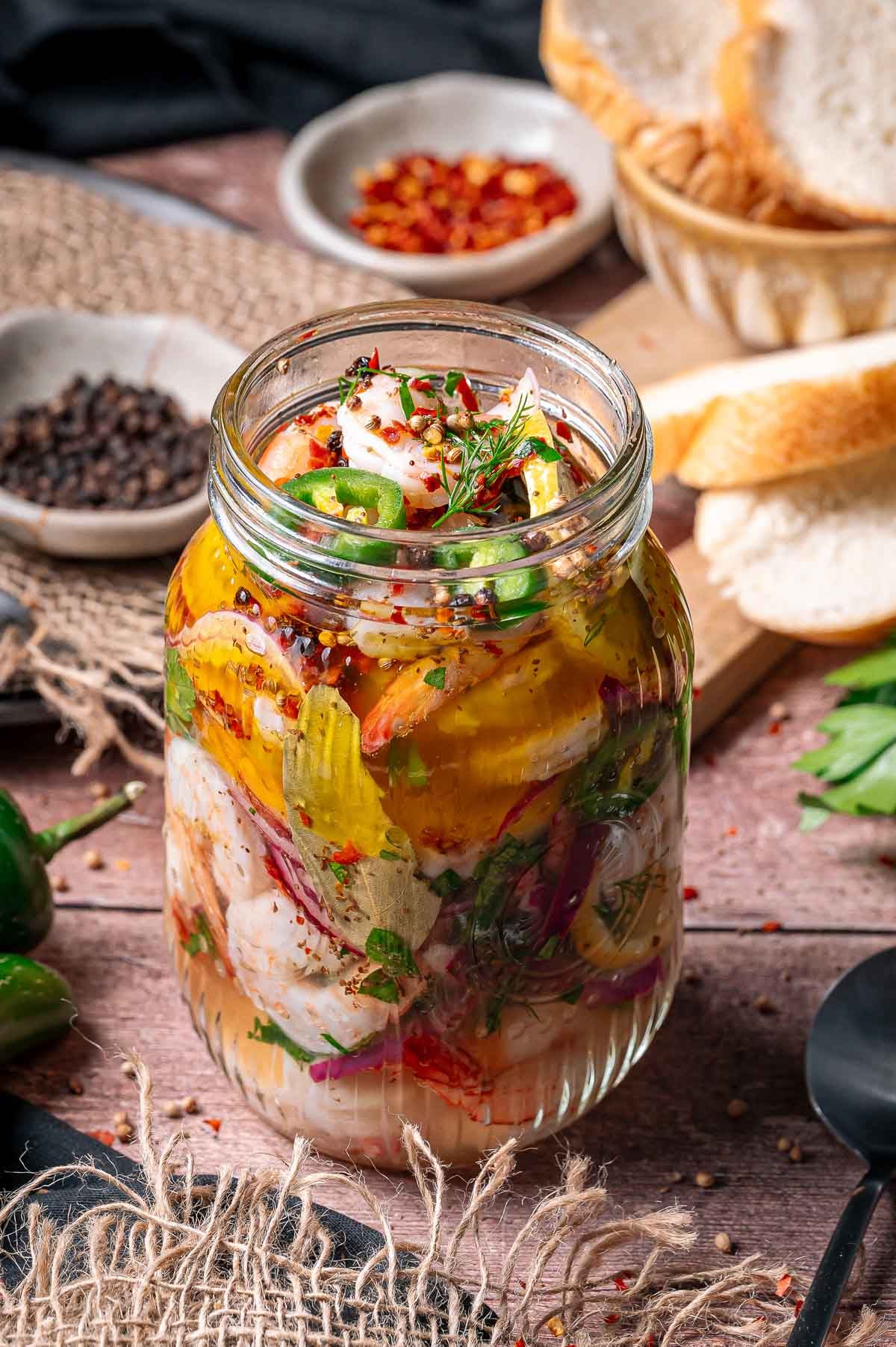 A glass jar filled with pickled shrim and herbs, surrounded by bread slices, spices, and fresh green peppers on a wooden table.