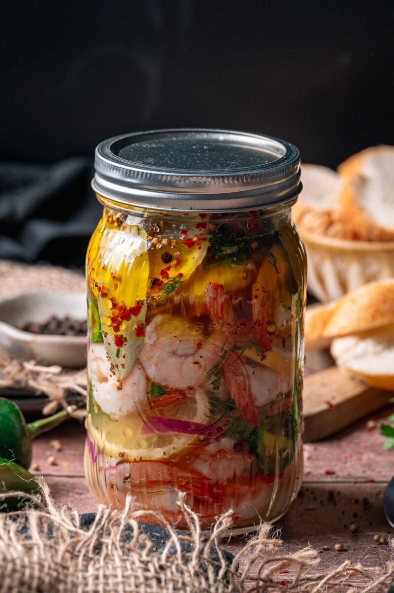 A glass jar filled with pickled shrimp, herbs, and spices sits on a table next to sliced bread and a small bowl of peppercorns.