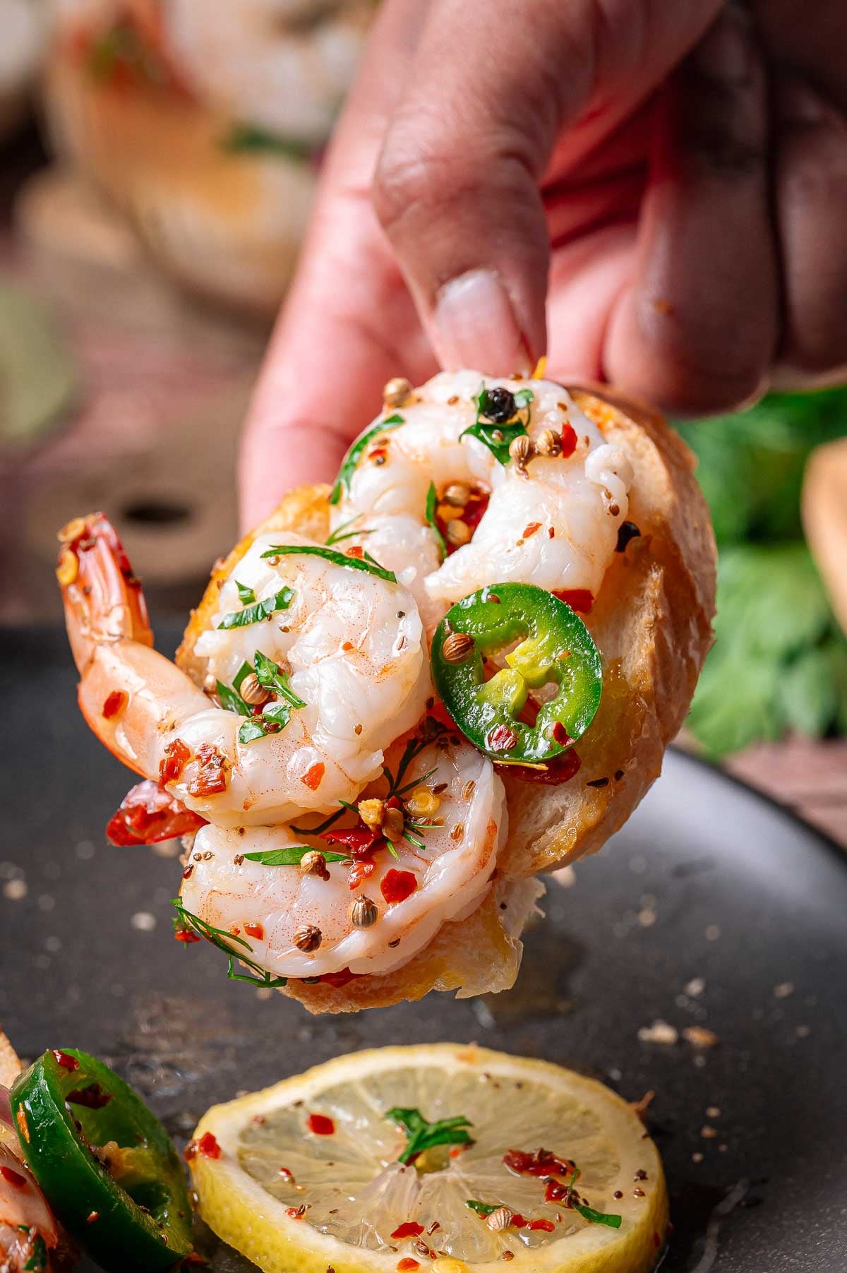 A hand holds a slice of toasted bread topped with pickled shrimp, sliced jalapeño, chopped herbs, and red pepper flakes. Lemon slices are visible on the plate below.