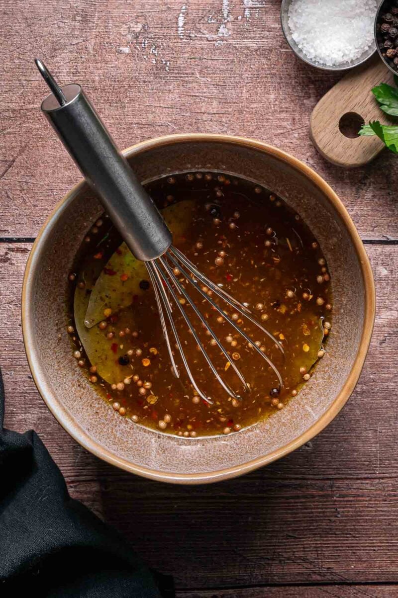 A bowl of pickle brine with visible spices and a metal whisk inside, placed on a wooden surface next to a small bowl of salt and herbs.