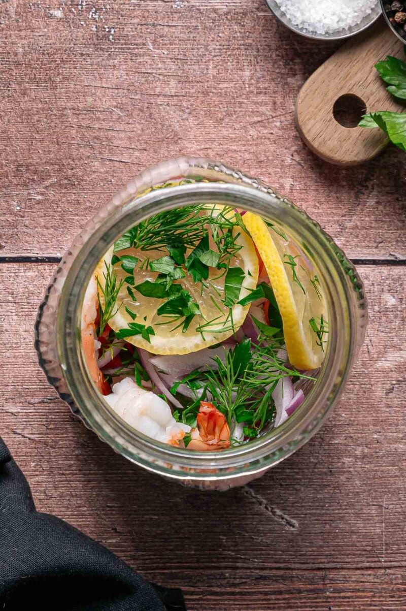 Overhead view of a glass jar filled with shrimp, lemon slices, red onion, fresh dill, and parsley on a wooden surface.