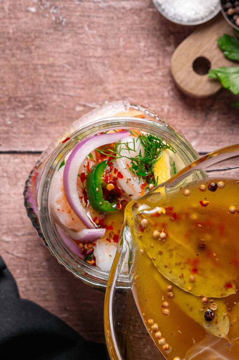 Brine is being poured from a glass jar over shrimp, onion slices, dill, lemon, and spices in a glass container on a wooden surface.