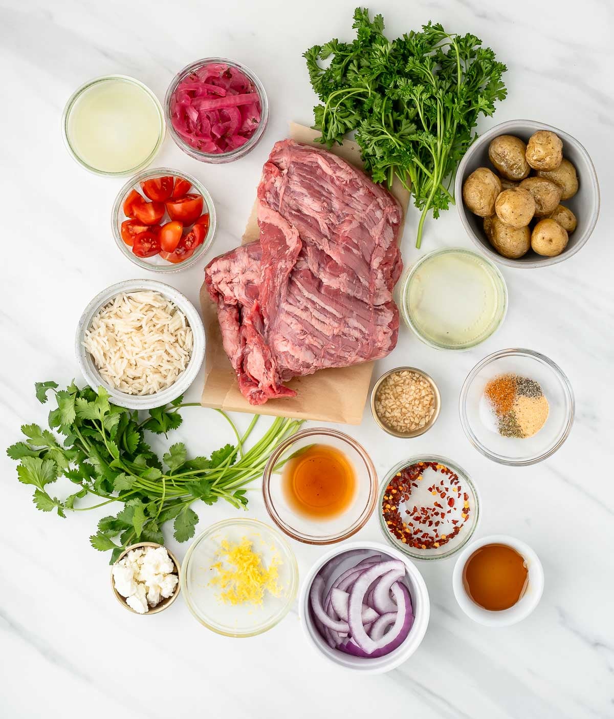 Ingredients for a beef bowl arranged on a white surface, including raw beef, potatoes, herbs, tomatoes, onions, rice, spices, and various liquids in small bowls.
