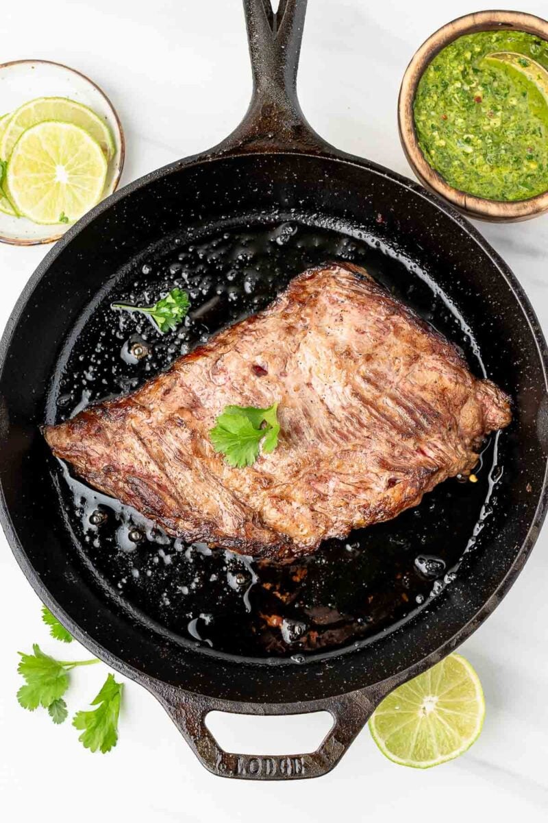 A cooked skirt steak sits in a cast iron skillet, garnished with cilantro, with a bowl of green sauce and slices of lime nearby.